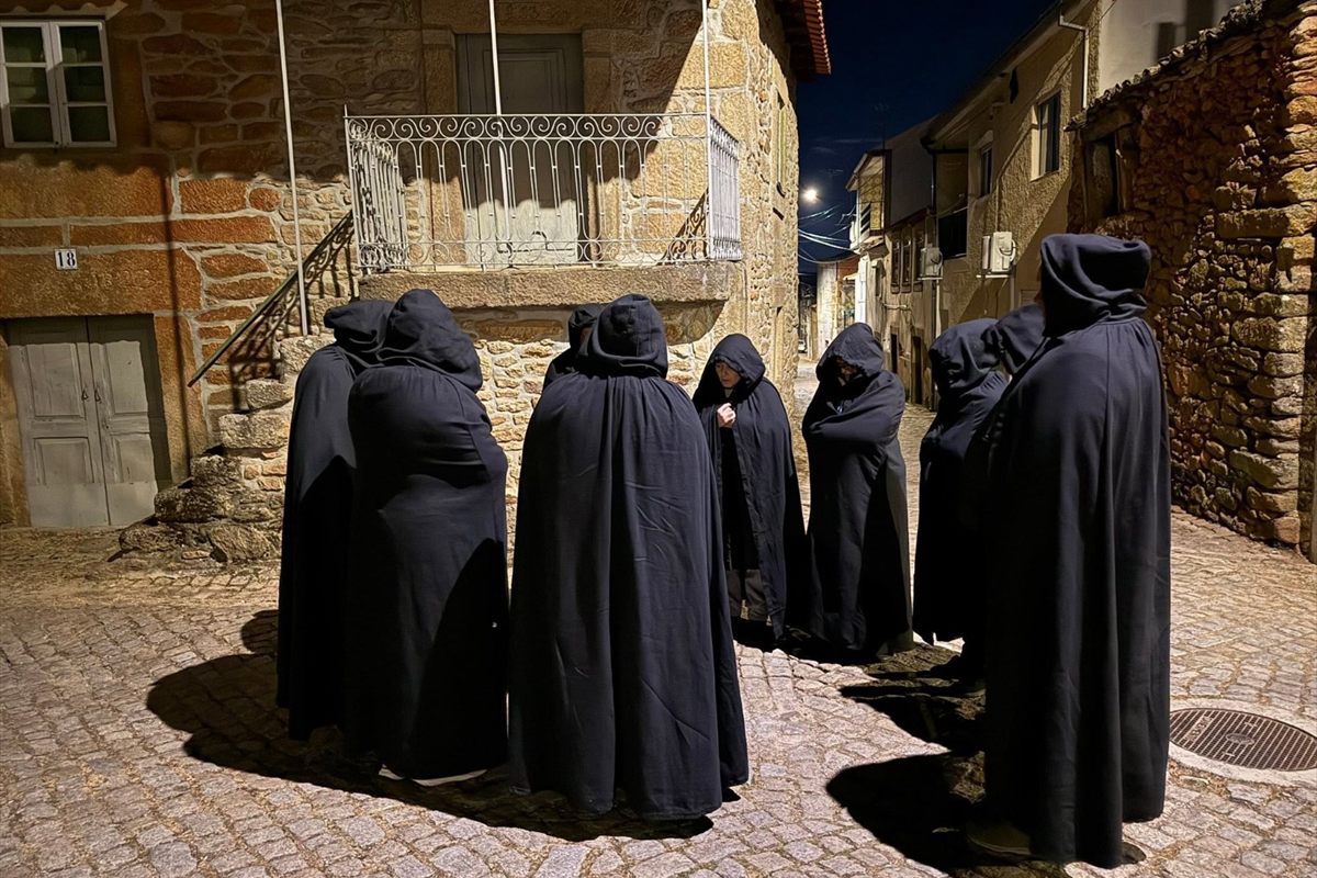 Grupo de figuras encapuzadas, de capa preta, numa rua de calçada da aldeia à noite, durante a Encomendação das Almas